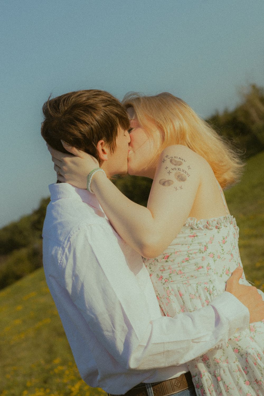 A couple shares a romantic kiss outdoors in a sunny field, she in a floral dress, he in a white shirt.