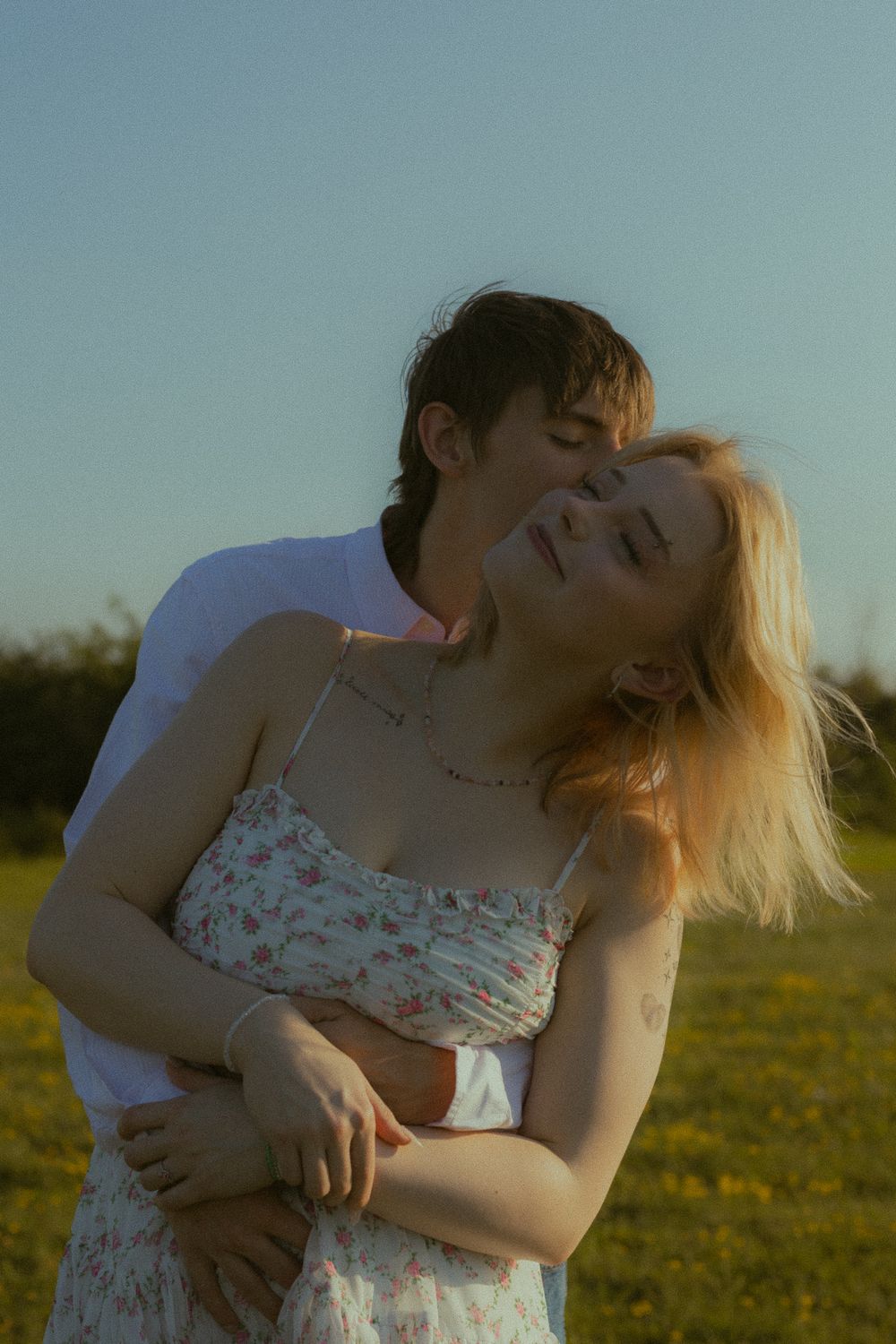 A couple embraces in a sunny field, the blonde woman in a floral dress tilting her head back with eyes closed.