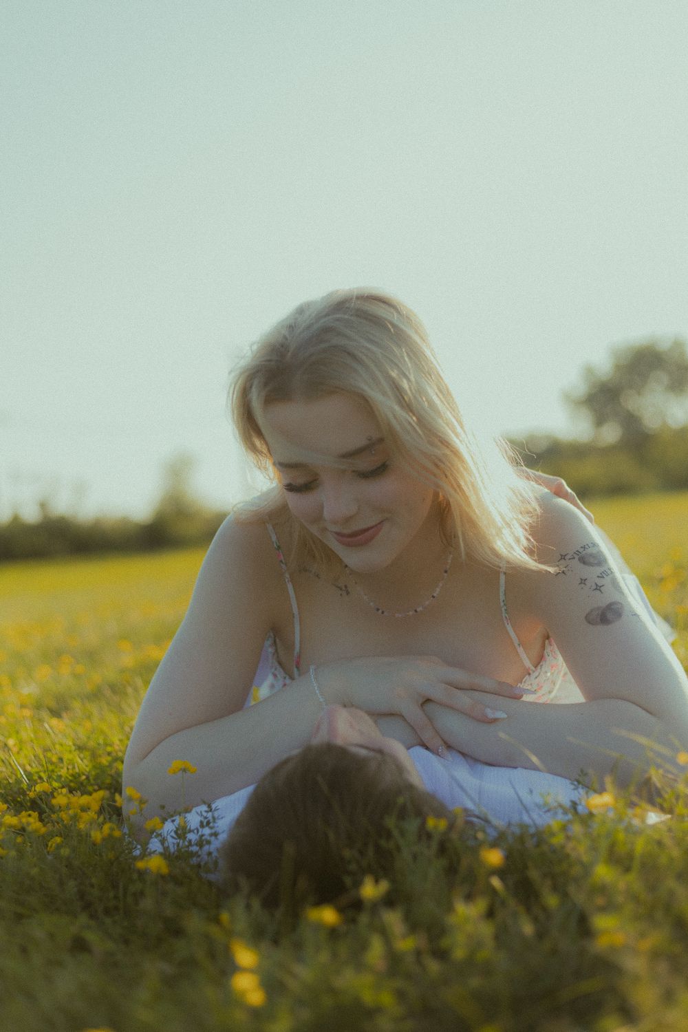 A blonde woman with tattoos lies in a sunny wildflower field, smiling down at a baby she is nursing.