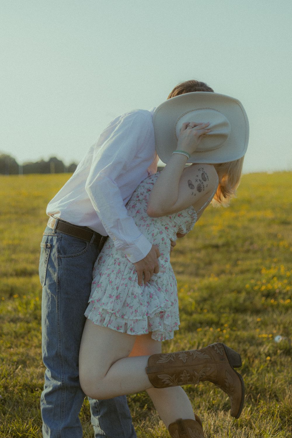 Couple sharing a romantic kiss in a sunny field, woman wearing a floral dress and cowboy boots, man holding a white hat.