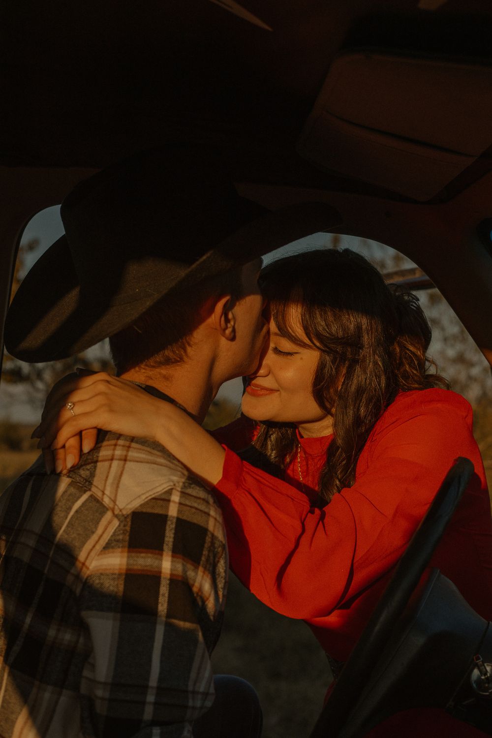 Intimate moment between couple in car with plaid shirt and red sweater during sunset.