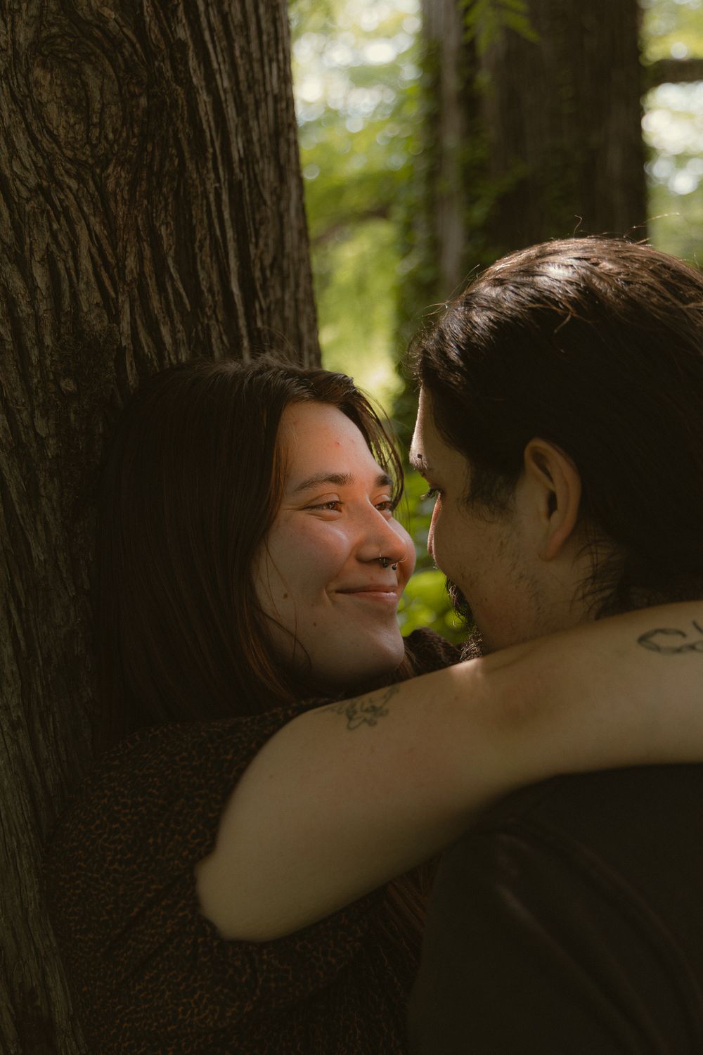 A couple shares a tender moment leaning against a tree in a sunlit forest, the woman smiling warmly.