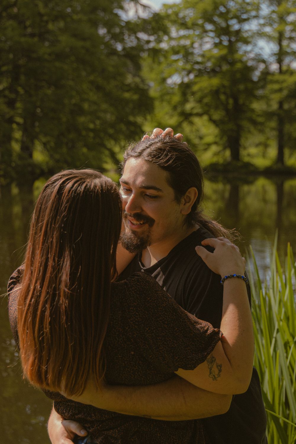 A couple embraces and smiles near a serene pond surrounded by lush green trees on a sunny day.