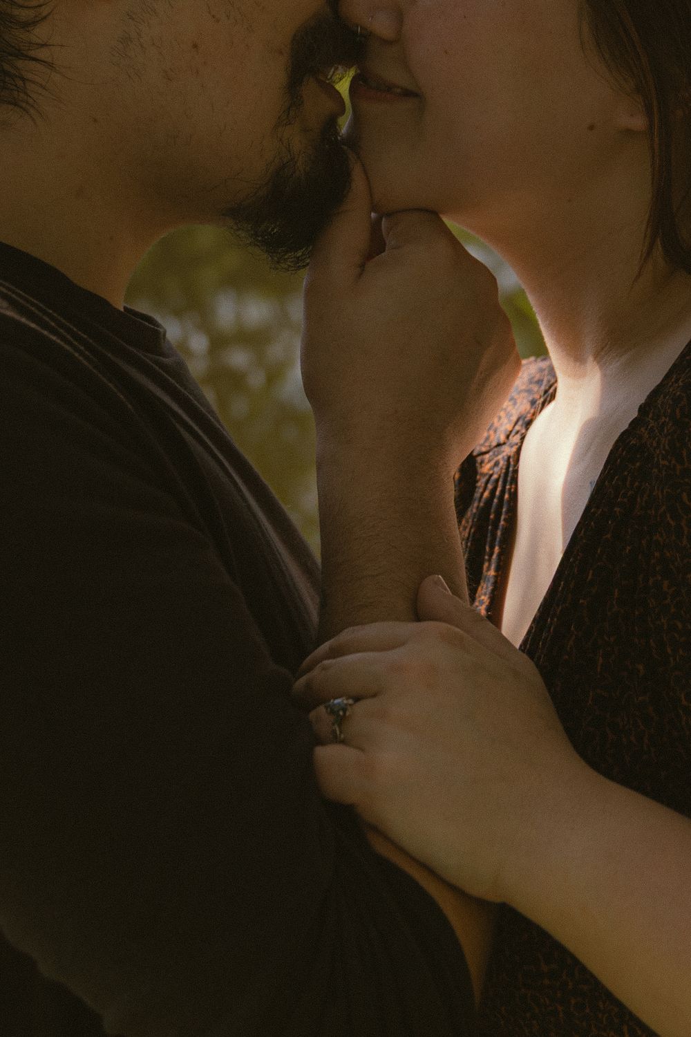 A couple shares a tender kiss outdoors, with a woman wearing a diamond ring holding her partner close.