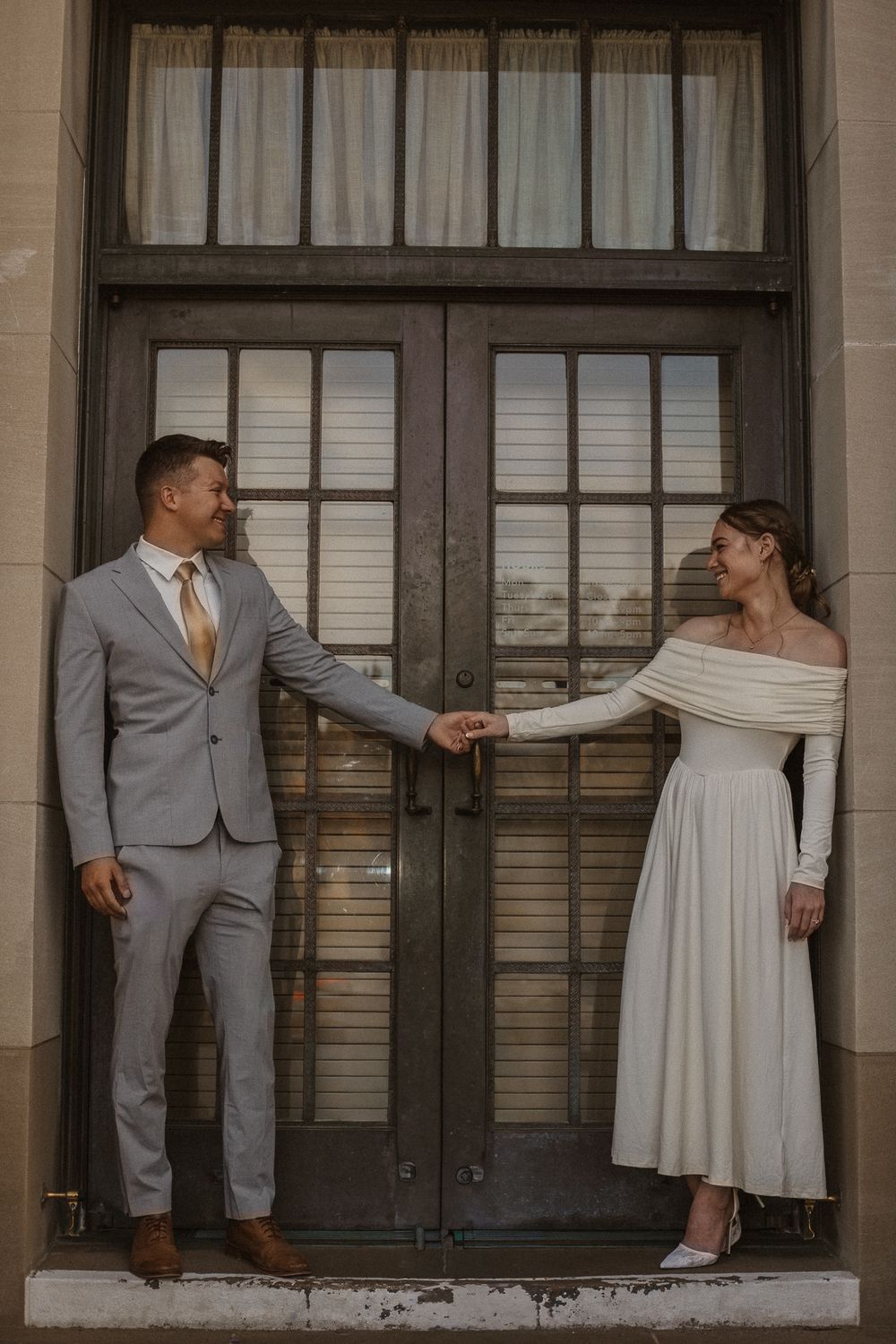 Couple in wedding attire poses playfully on a building porch with white columns.