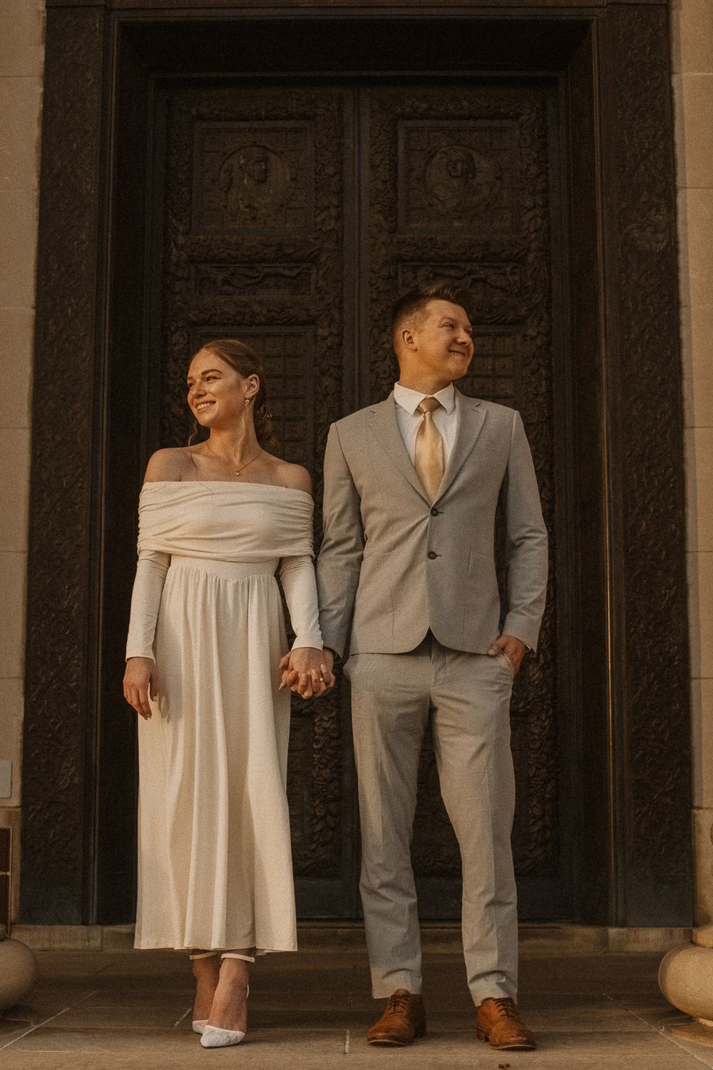 Bride in white dress and groom in gray suit pose together on building steps.