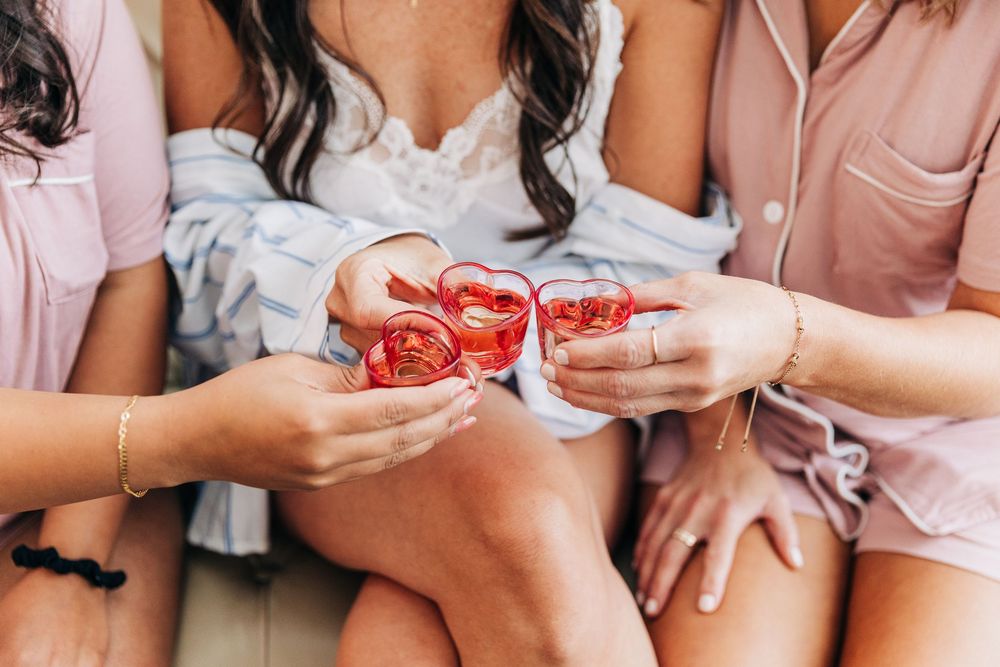 Row of people in pink and white outfits sitting on couch holding red tropical drinks during bridal party celebration.