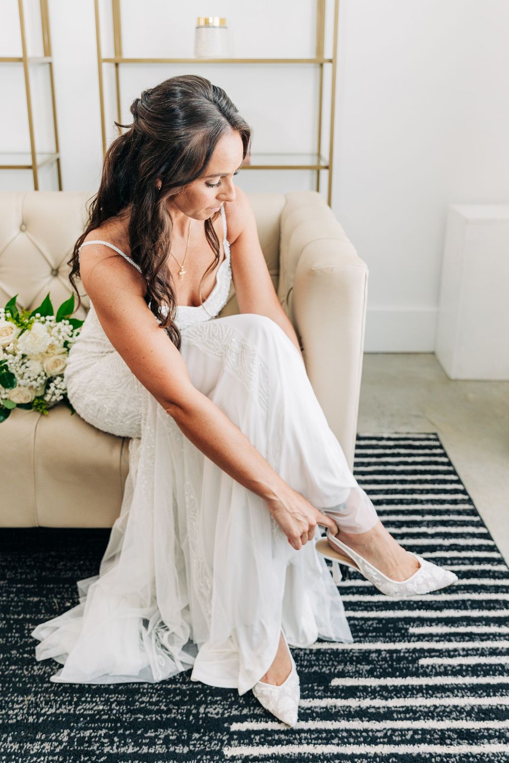 A person in a flowing white wedding dress sits on a beige armchair, adjusting elegant white heels on a striped rug.
