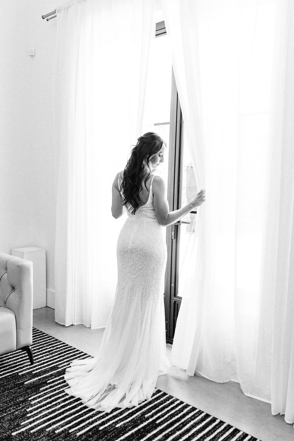 A bride in a fitted lace wedding gown stands by white curtains on a striped rug in black and white.