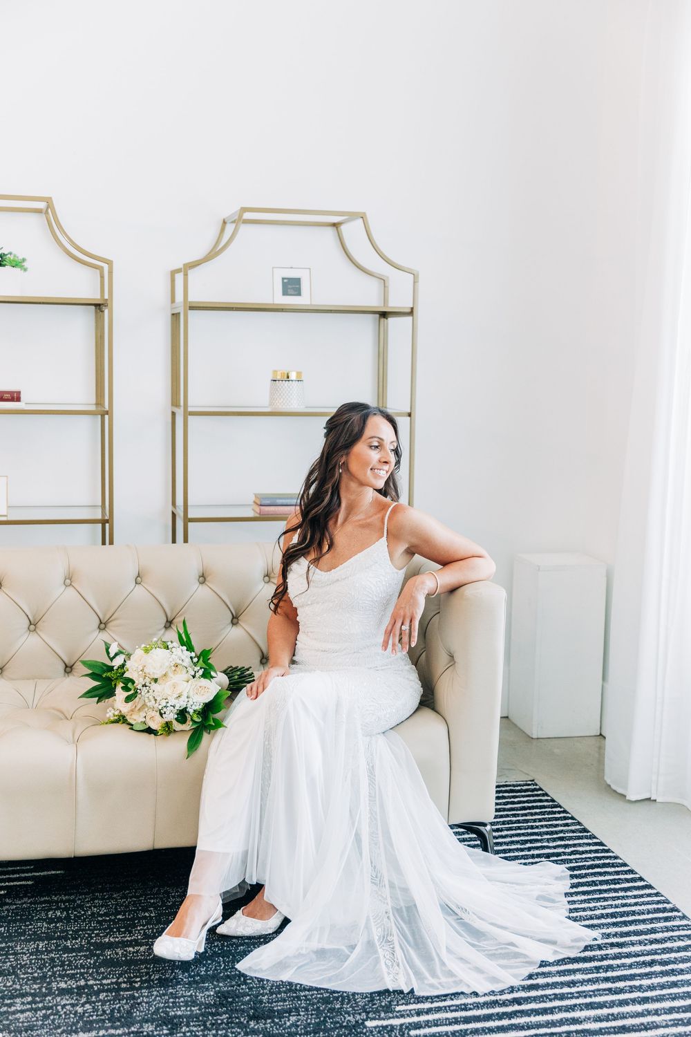 A figure in a white wedding dress relaxes on a beige chaise lounge with white floral decorations against a neutral wall.