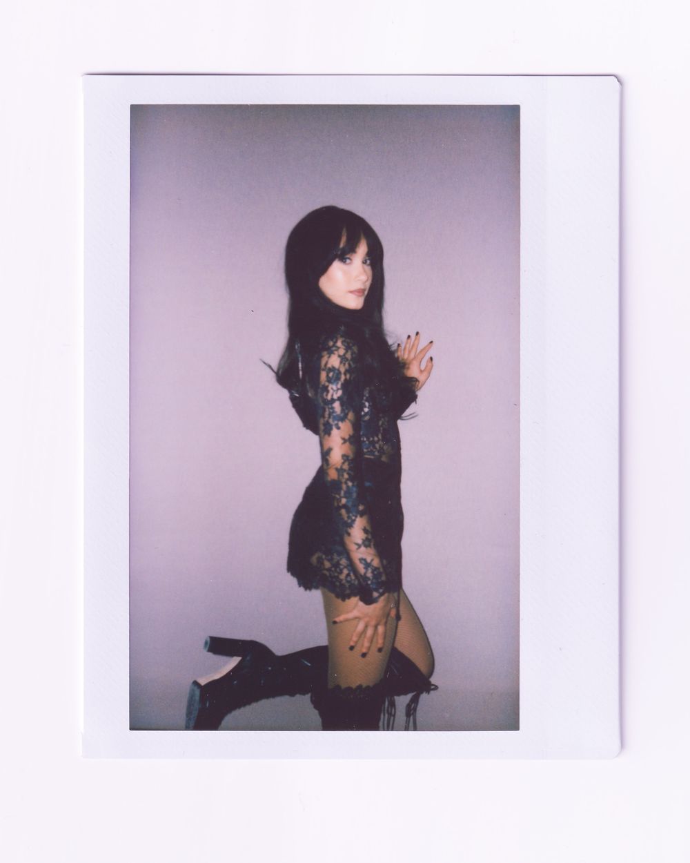 Woman in black lace mini dress and thigh-high boots poses for a Polaroid-style photo against a purple background.