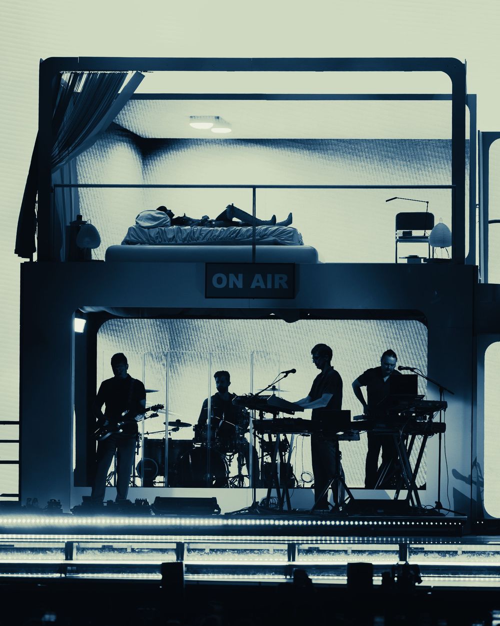 Silhouetted band performing on stage beneath an ON AIR sign, with a person lying on a platform above them.
