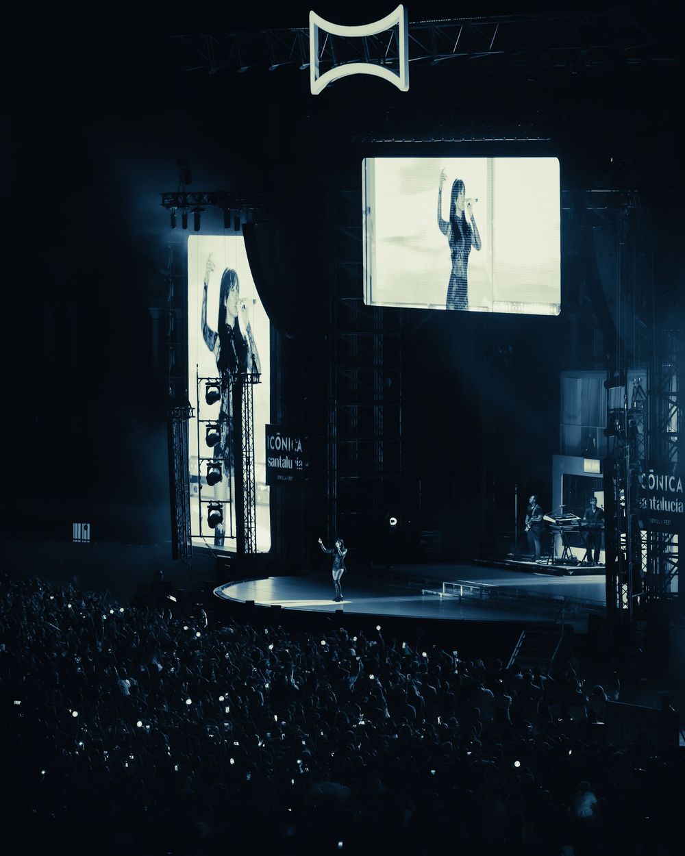 A performer on a concert stage with large screens displaying the artist to a massive crowd in a dark arena.