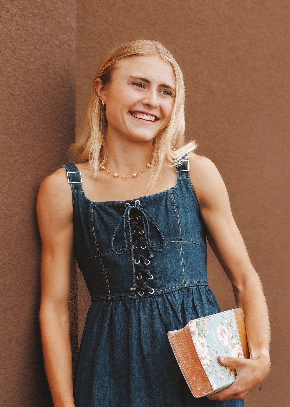Smiling blonde woman in a denim lace-up dress holding a floral book against a warm adobe wall.