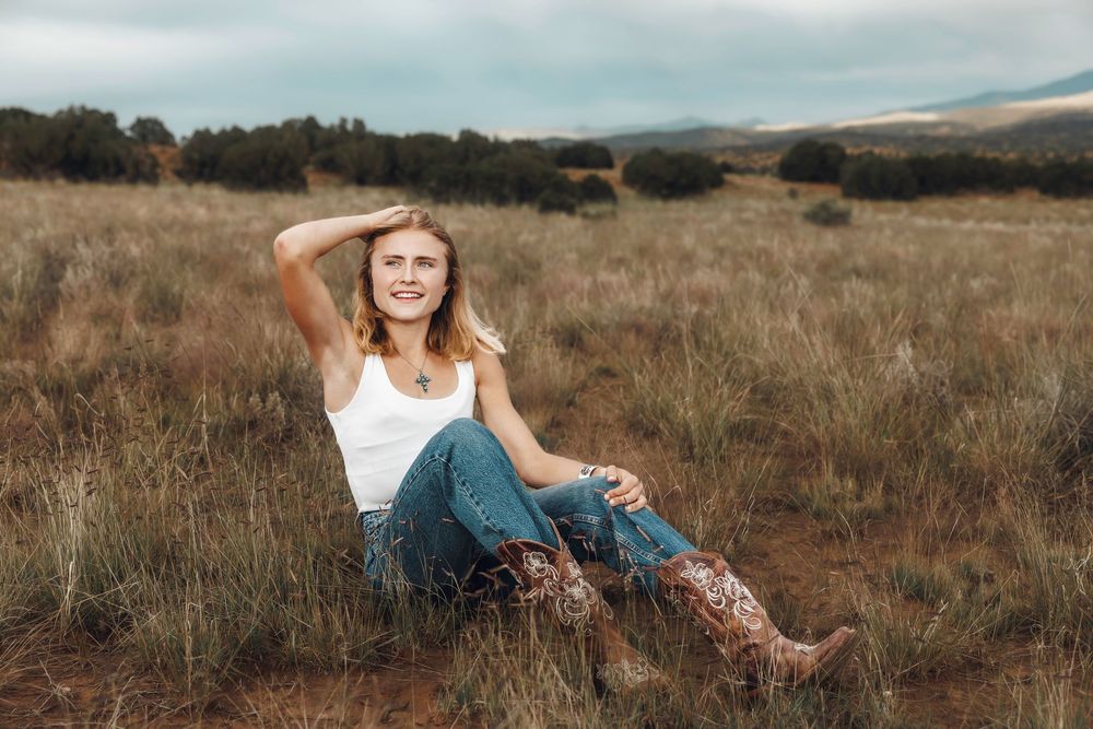 Young blonde woman in white tank top, jeans, and cowboy boots sitting in a grassy field under cloudy skies.