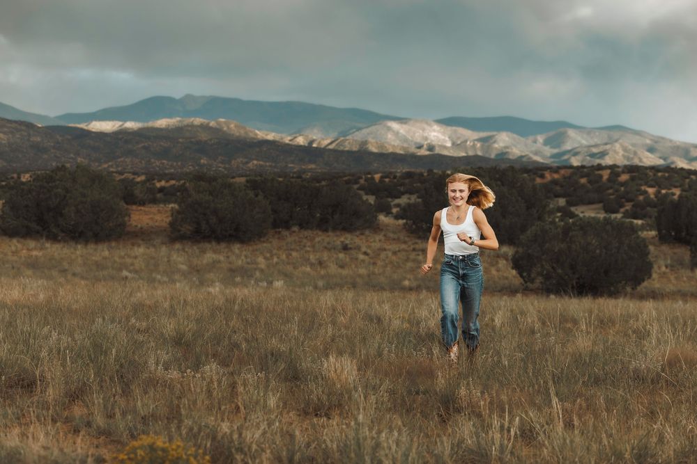 Young blonde woman in white tank top and jeans running freely through a wide open field near mountains.