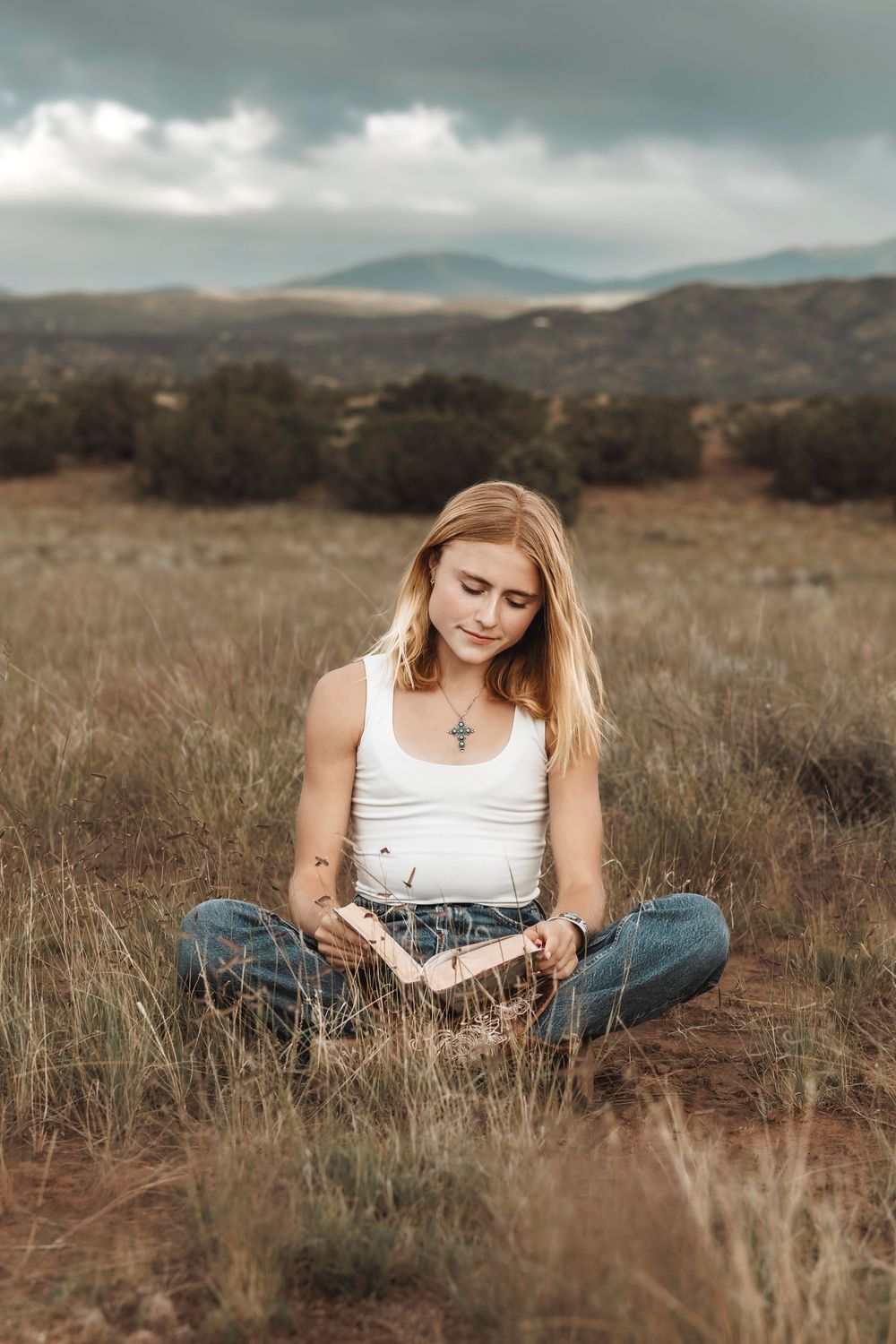 Young woman with cross necklace reading a book while sitting cross-legged in a scenic open field.