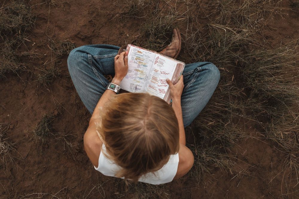 Overhead view of a woman in jeans and cowboy boots reading a book while sitting on dry ground.