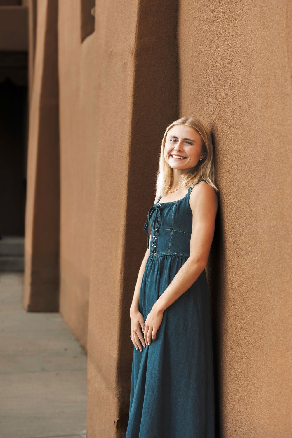 Smiling blonde woman in a teal sundress leaning against a warm adobe wall in soft natural light.
