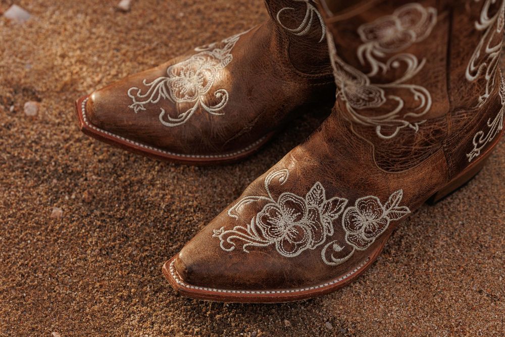 Close-up of brown leather cowboy boots with white floral embroidery resting on sandy desert ground.