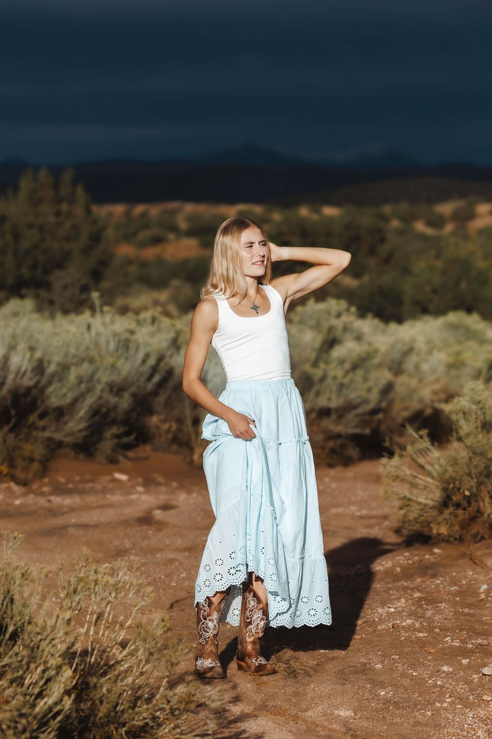 Blonde woman in a white tank top and light blue maxi skirt with cowboy boots on a desert trail.