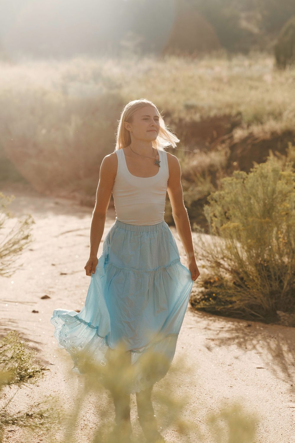 Blonde woman in a white tank top and flowing blue skirt walking on a sandy desert trail at sunset.