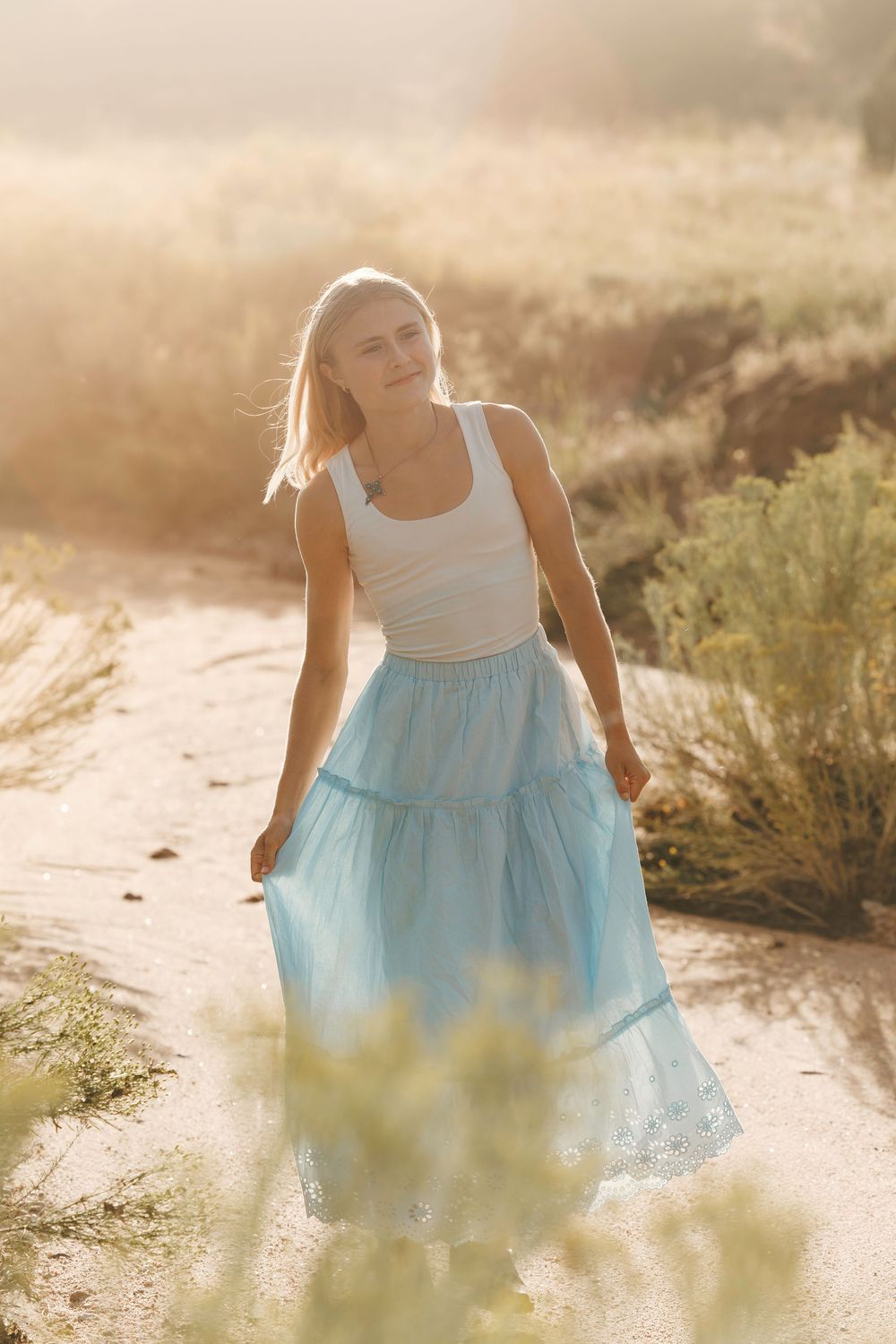 Blonde woman in a white tank and blue tiered maxi skirt walking through a sun-drenched desert path.