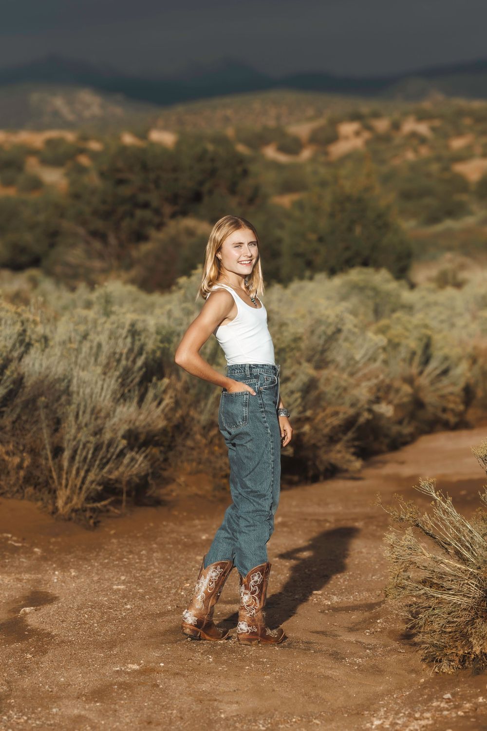 Smiling blonde woman in white tank, jeans, and embroidered cowboy boots posing in a desert landscape.