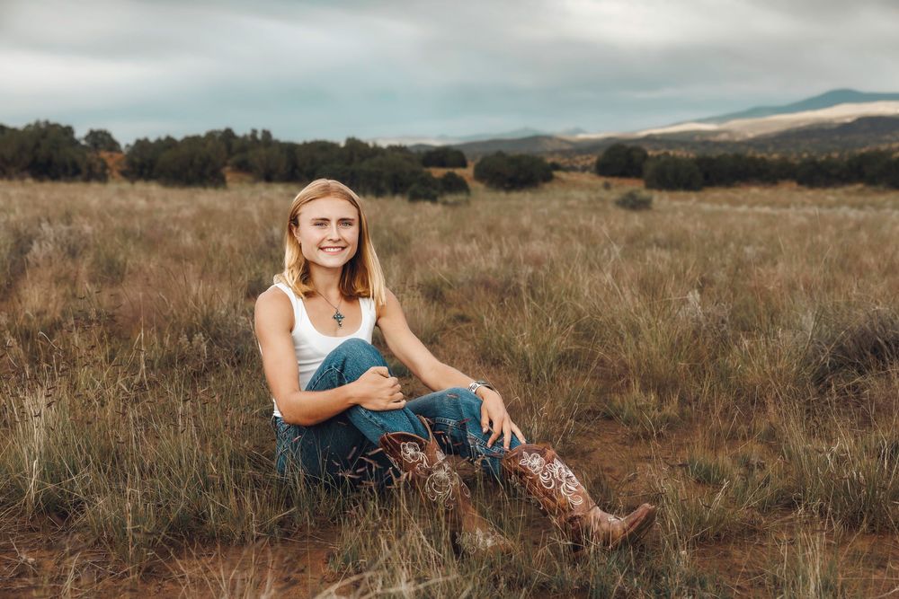 Smiling blonde woman in white tank and jeans with floral cowboy boots seated in a rustic open field.