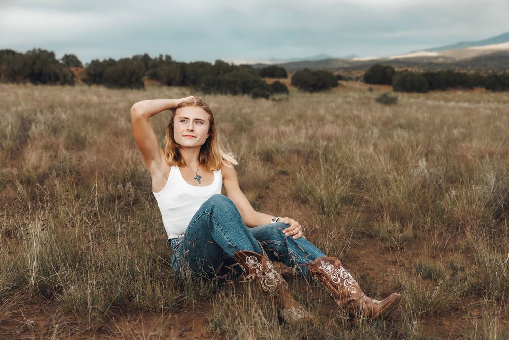 Blonde woman in white tank top and jeans relaxing in a dry grass field with mountains in the distance.