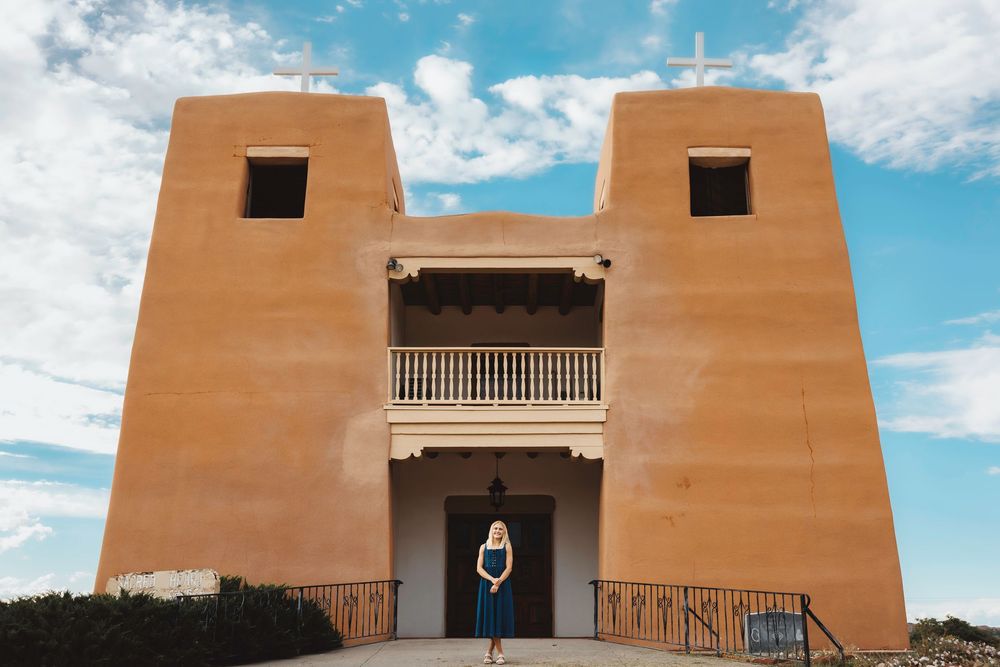 Santa Fe senior standing in the arched doorway of a historic adobe church under a blue sky in New Mexico.