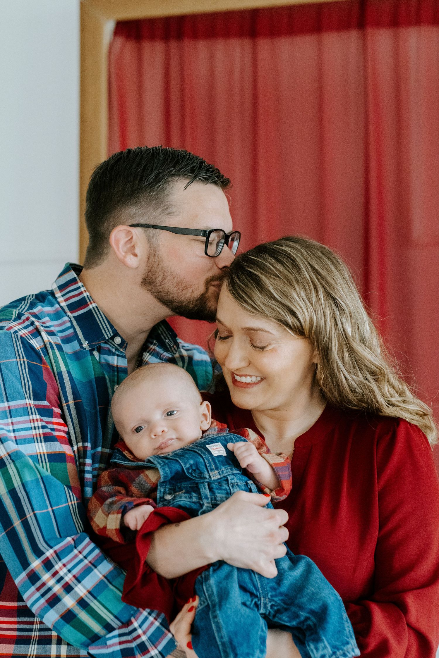 Family of three in coordinating red and plaid outfits share loving embraces during indoor photo session.