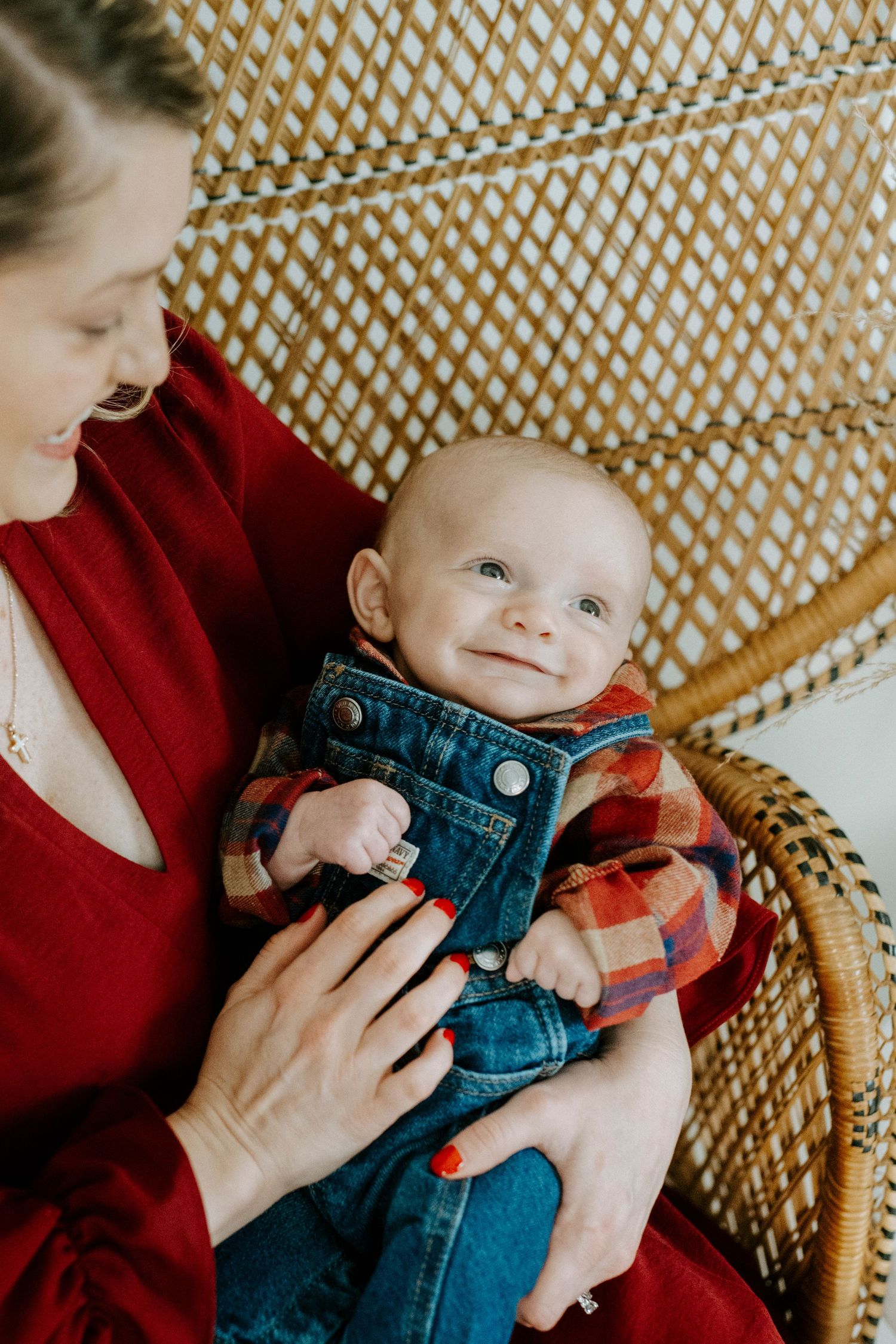 A baby in denim overalls and plaid shirt sits in a wicker chair while being held by someone in red.