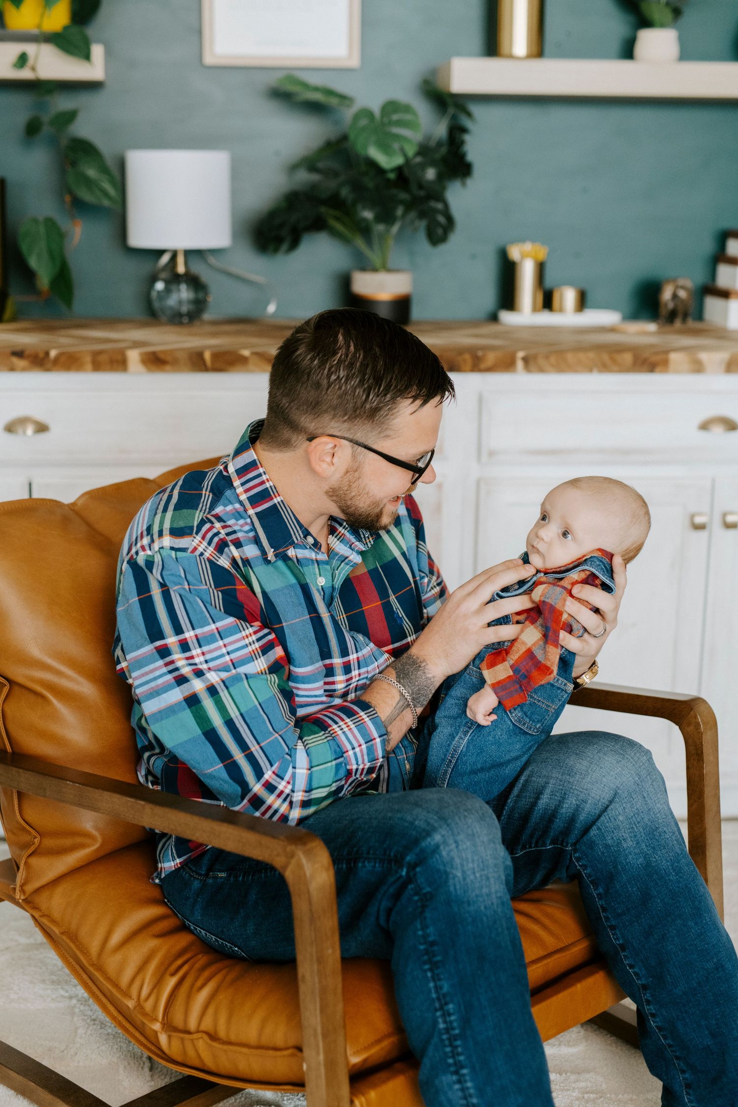 Adult in plaid shirt sits on brown leather chair while holding and interacting with baby in casual home setting.