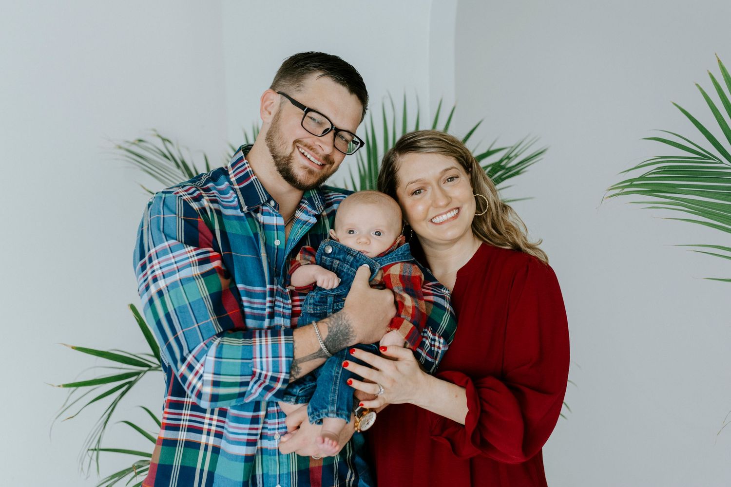 A family poses in coordinated red and plaid outfits against a white wall with tropical plants.
