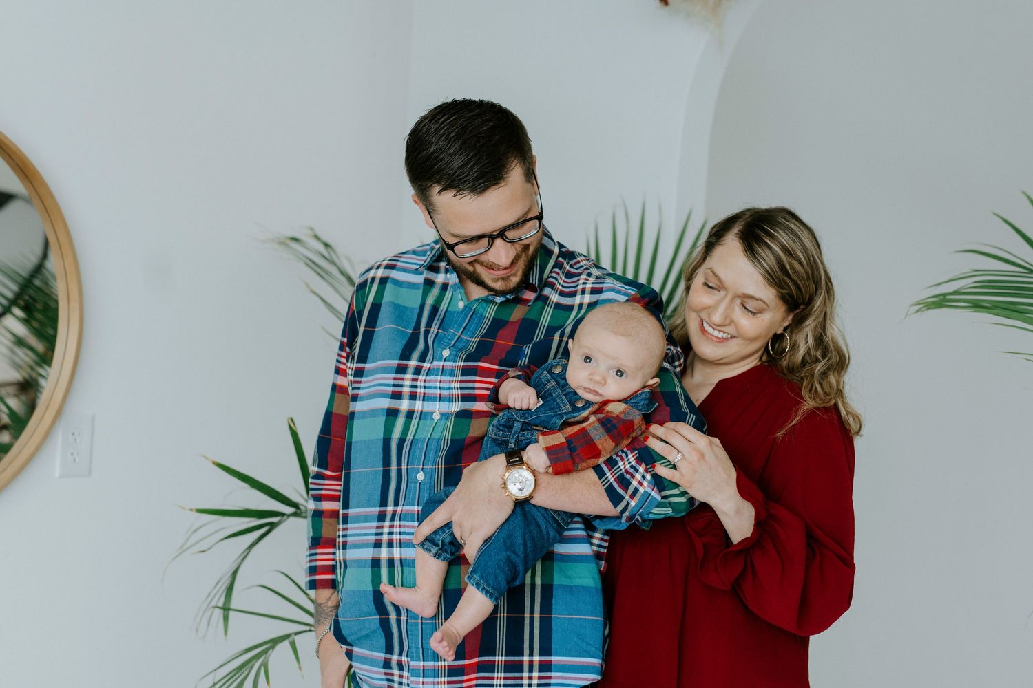 A couple in plaid and burgundy embraces warmly while standing against a white wall with tropical plants.