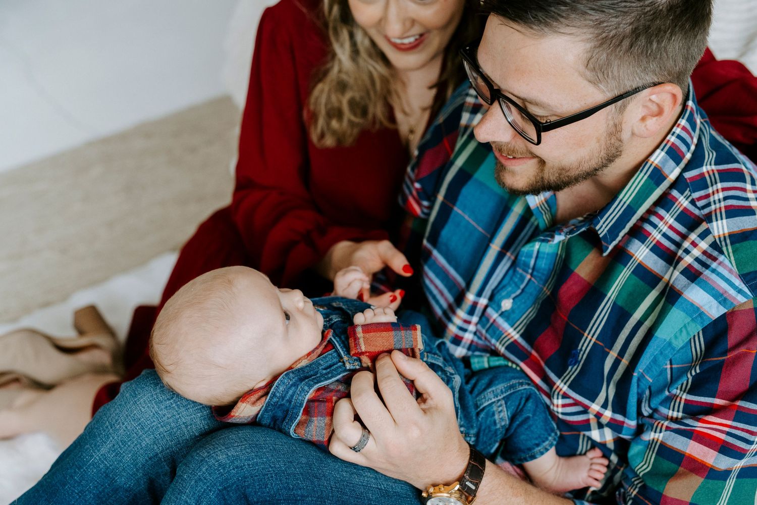 Parents in red and plaid clothing share a tender moment with their baby on a light colored floor.