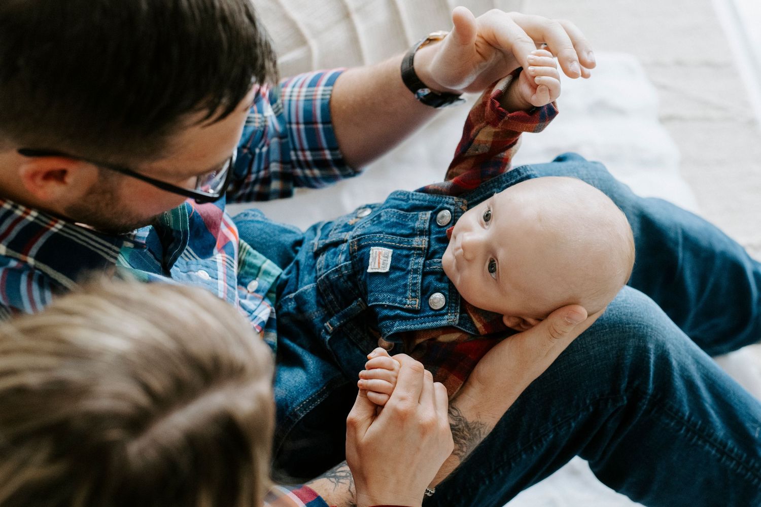 Two people in casual clothing tenderly care for a baby wearing blue overalls on a soft white surface.