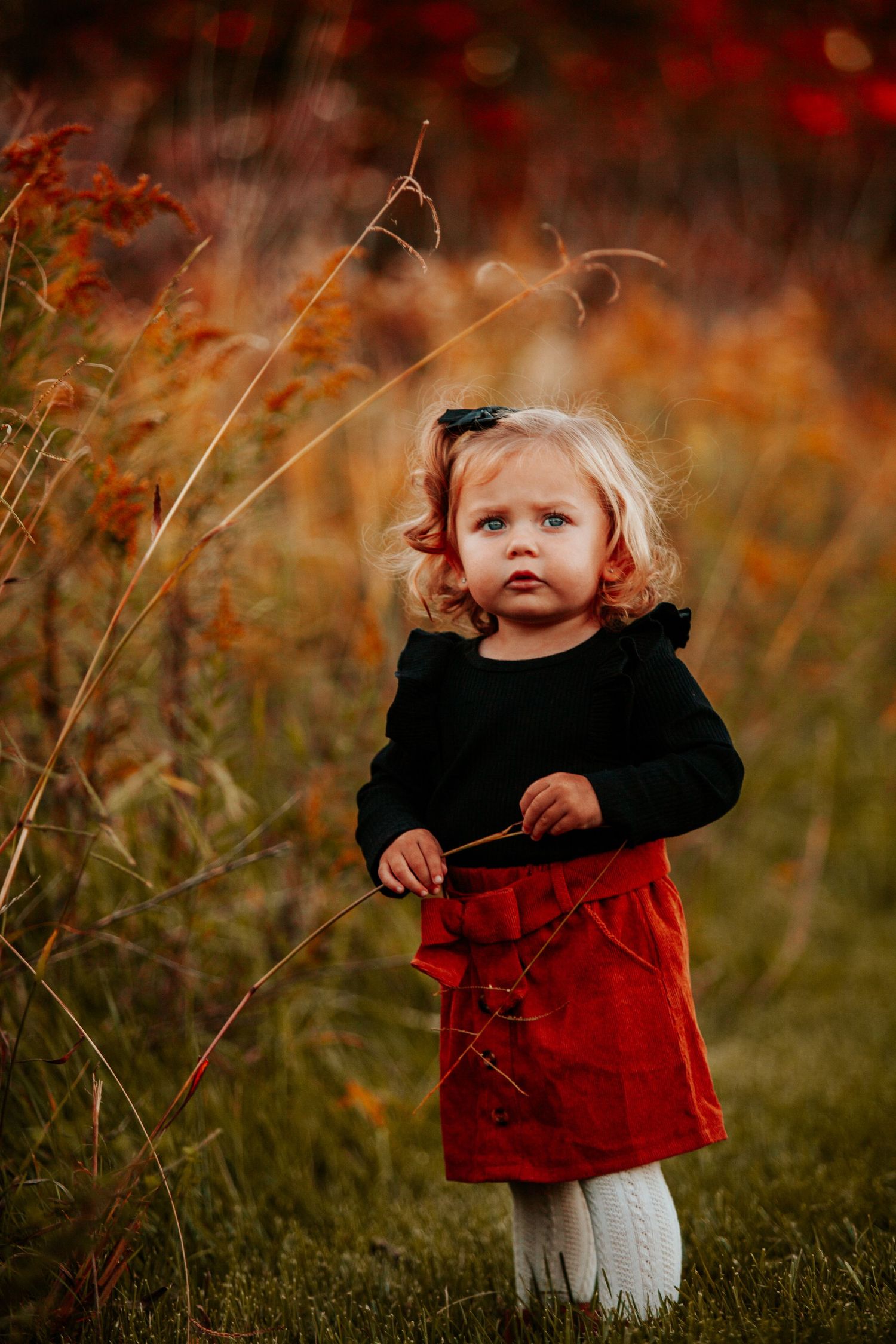 A young child in a black top and red skirt plays among tall grasses at sunset.