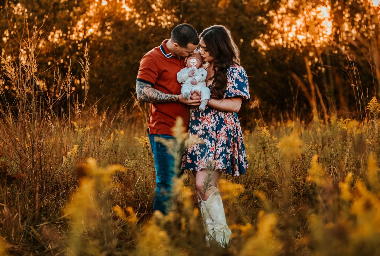 A couple holds their baby while standing in a field of yellow wildflowers at sunset.
