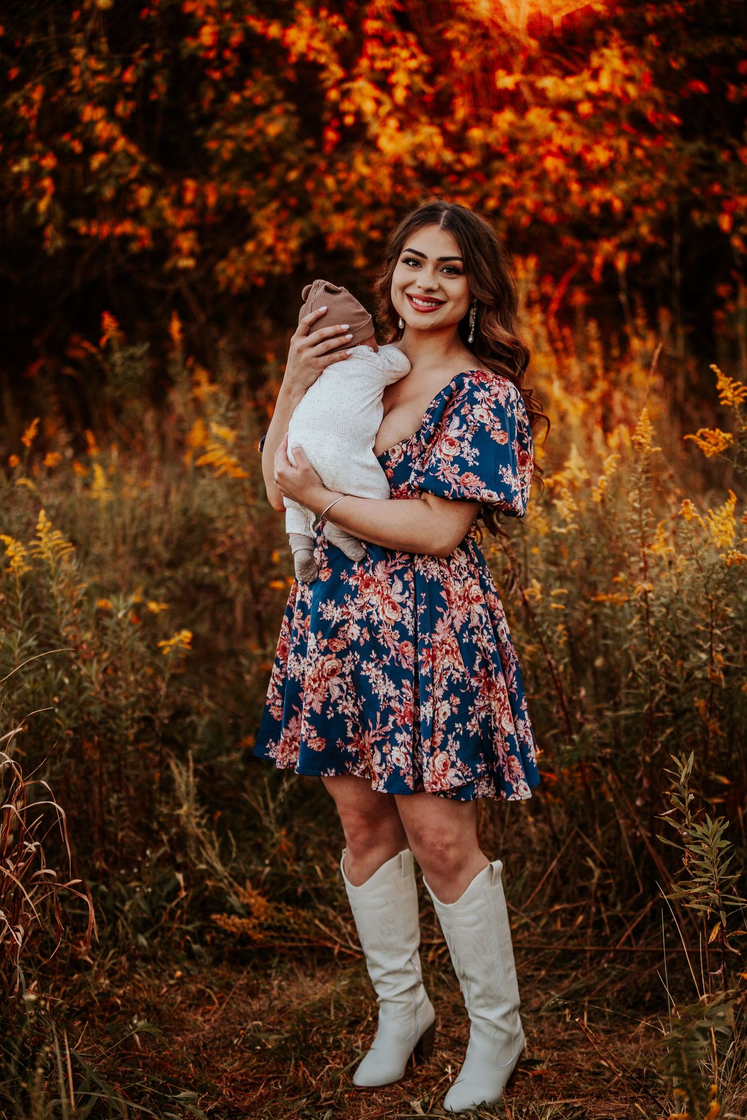 A person in a floral dress and white boots holds a baby against an autumn backdrop.