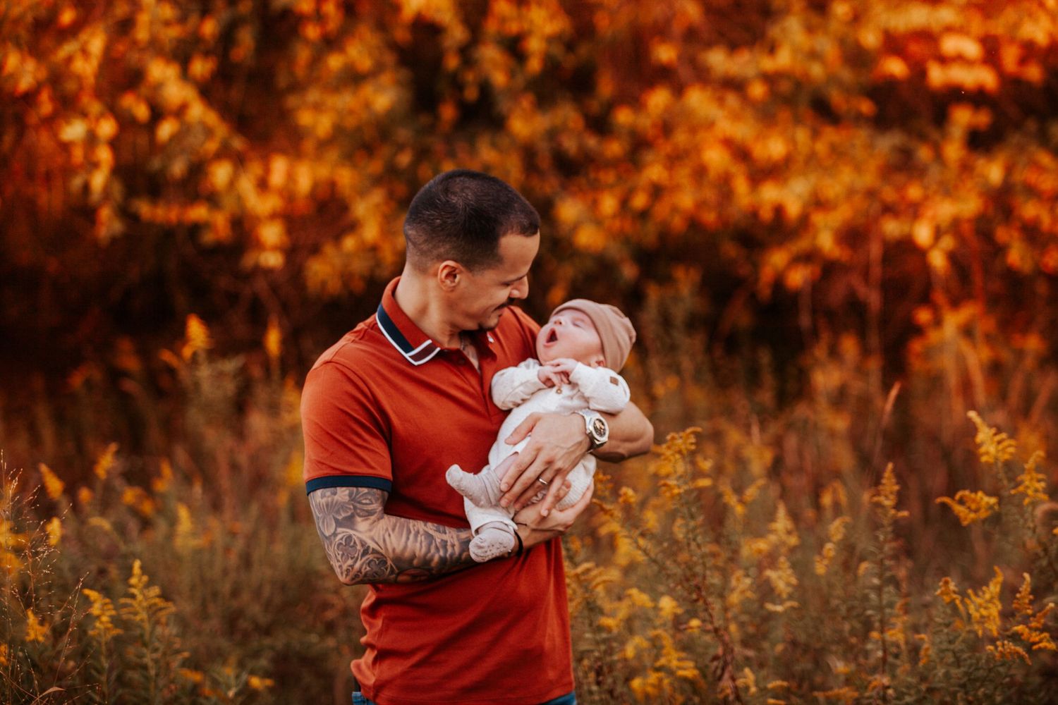 A person in a red polo shirt holds and gazes at a baby in a golden sunset field.