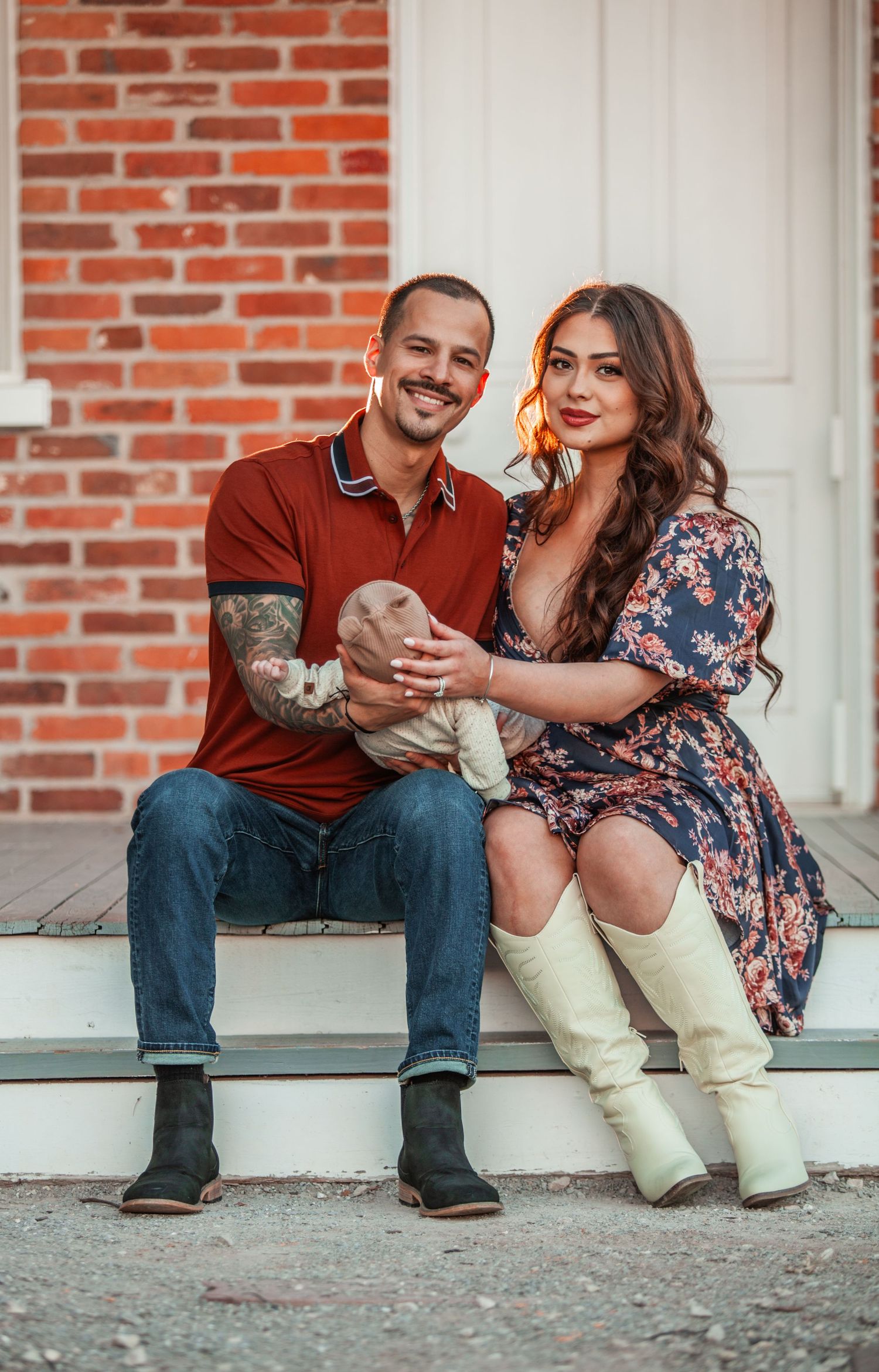 A couple sits on brick steps with their baby, wearing casual attire and boots.