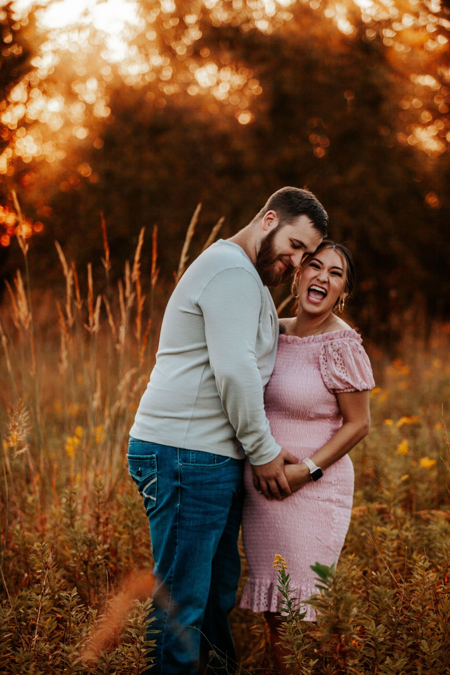 A couple shares a joyful moment in a field at sunset with golden bokeh lighting in the background.