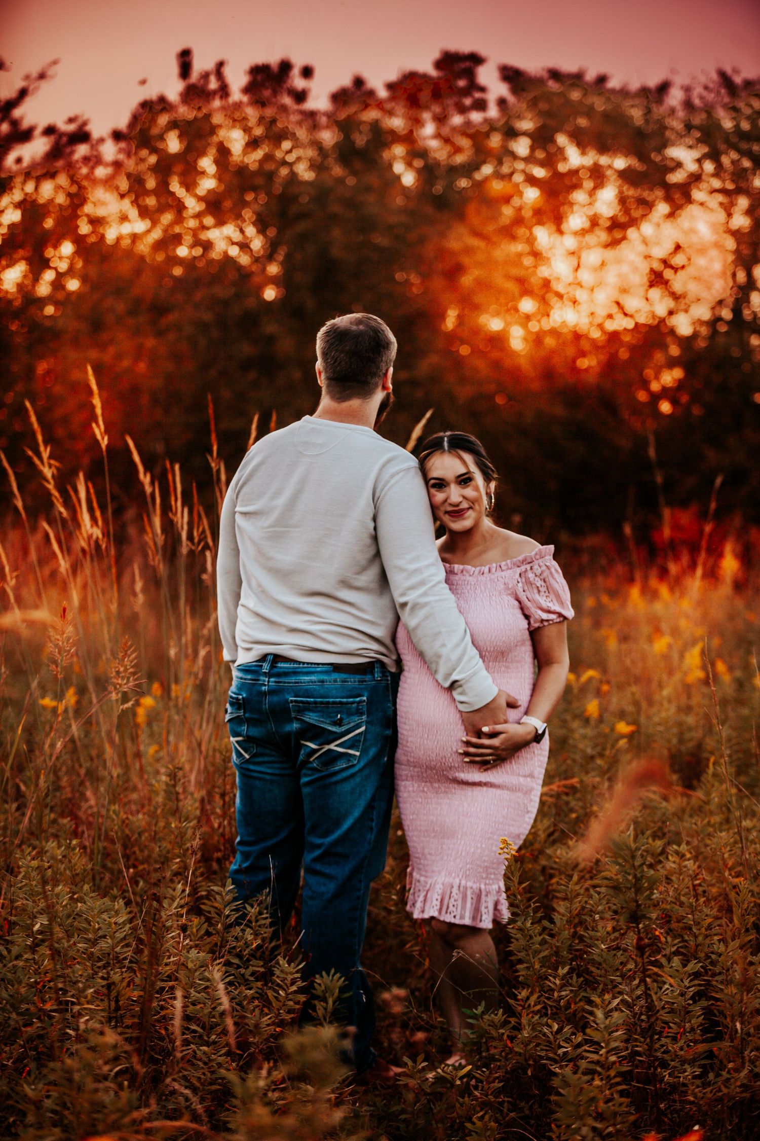 A couple stands together in a golden field during sunset wearing pink and cream colored outfits.