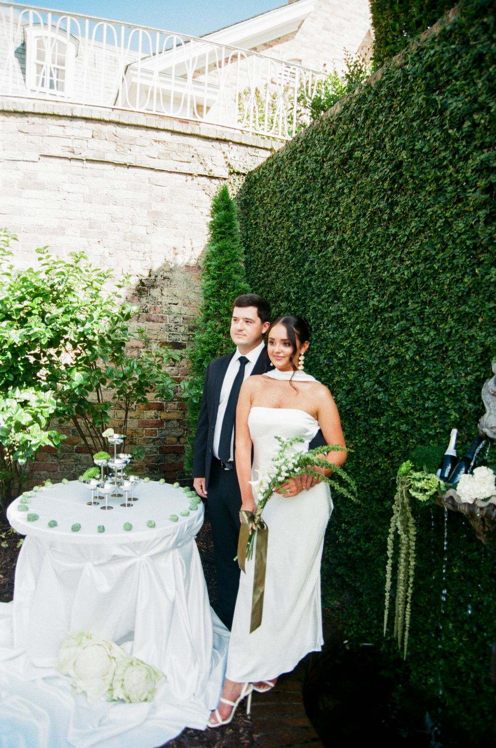 Bride and groom pose elegantly in front of a lush green hedge wall at an outdoor wedding reception.