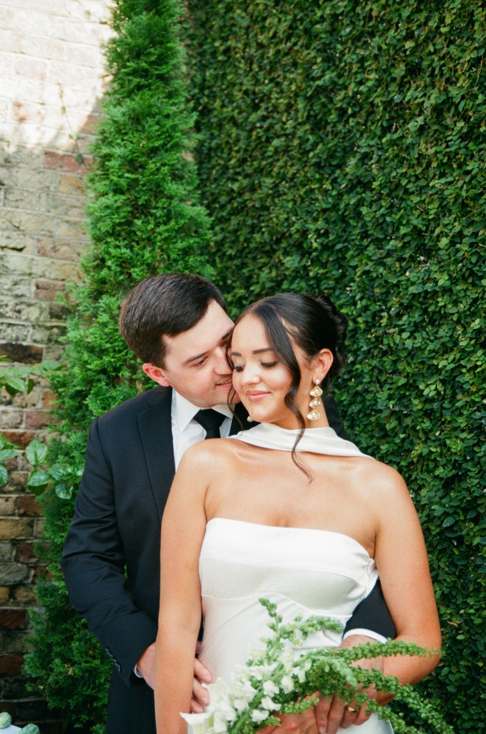 Romantic wedding moment captured against lush green hedge backdrop in garden setting.