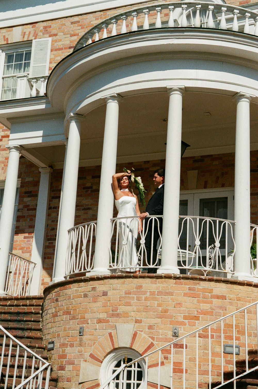 Wedding party stands on curved white balcony of elegant brick mansion with classical columns and architectural details.