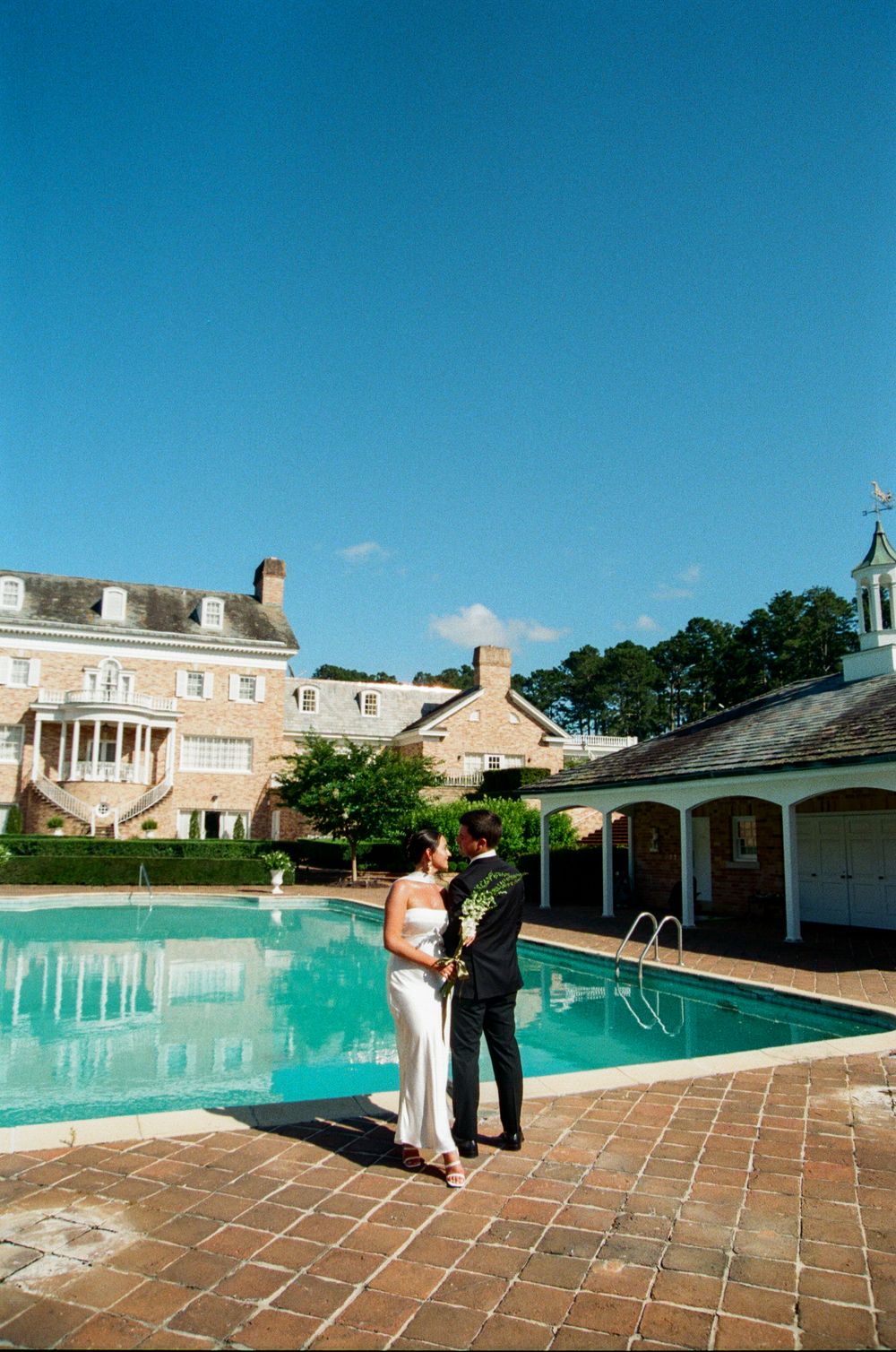 A serene swimming pool setting with tile patio and residential buildings in the background under a bright blue sky.