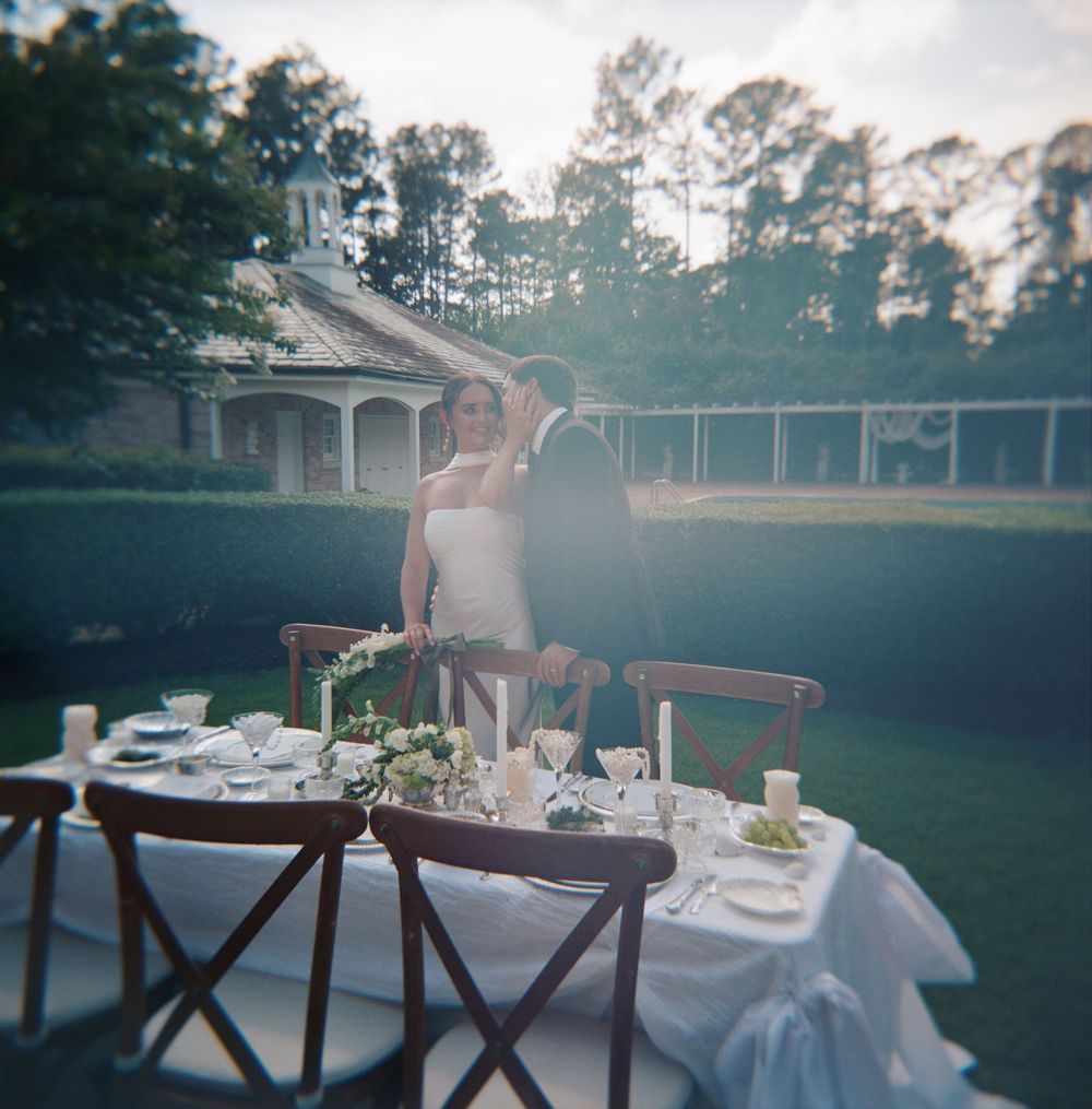 Two long dining tables with white cloths and chair settings arranged outdoors for an elegant garden party at sunset.